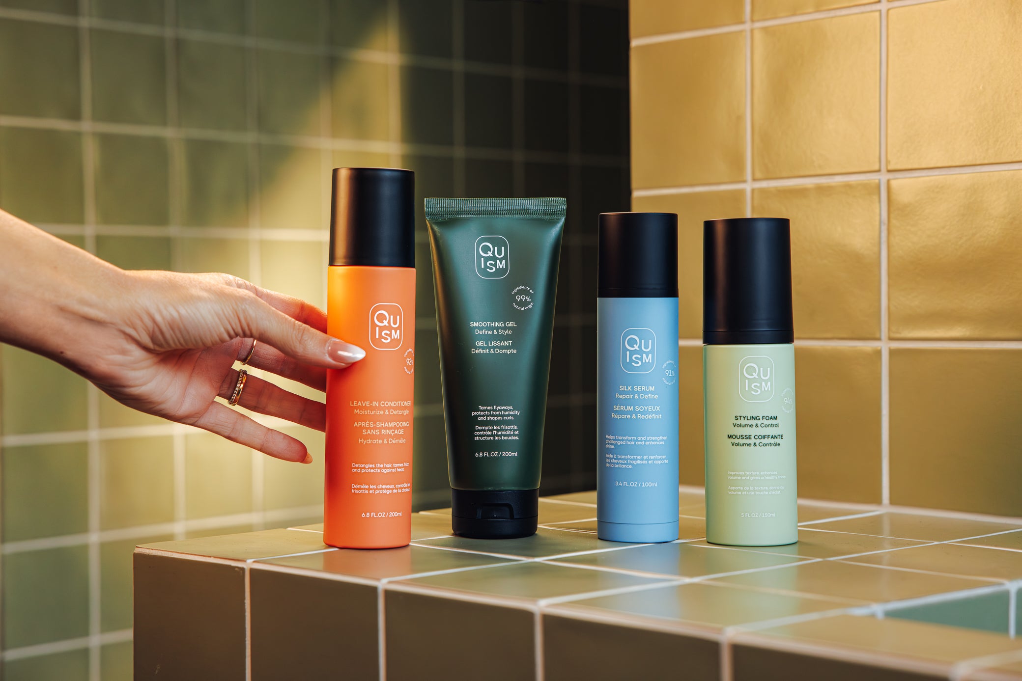 Four Quism haircare products on a tiled bathroom counter with a hand reaching for one of them.
