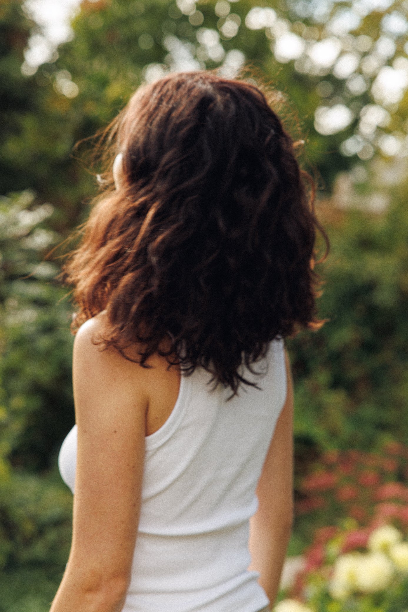 Person with wavy hair wearing a white tank top in a garden setting